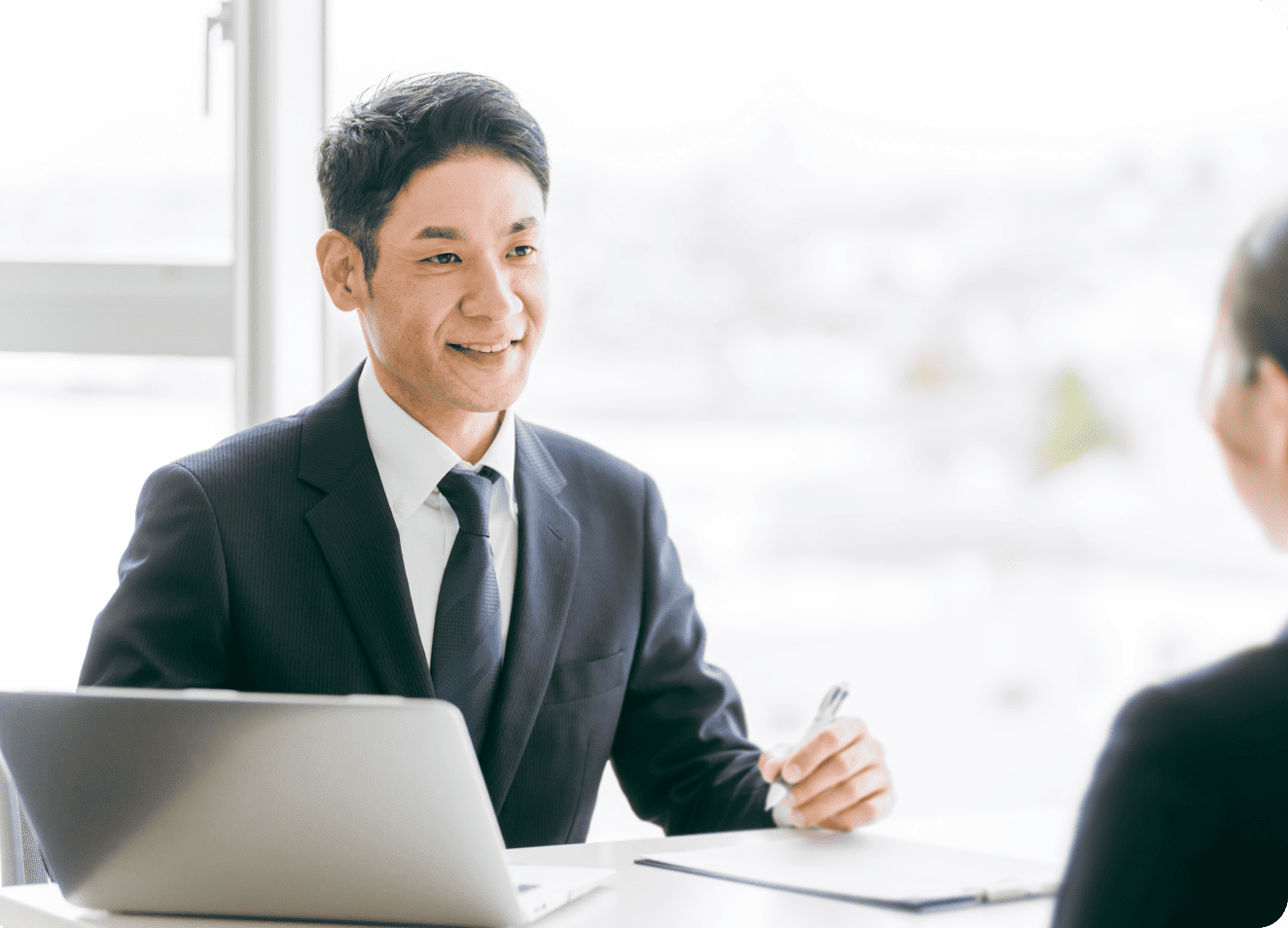 ノートパソコンのあるデスクで、笑顔のビジネスマンがペンを持ち、会議または面接をしている様子